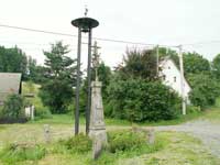 litinový křížek s kamenným podstavcem v Podolí, v pozadí zvonička